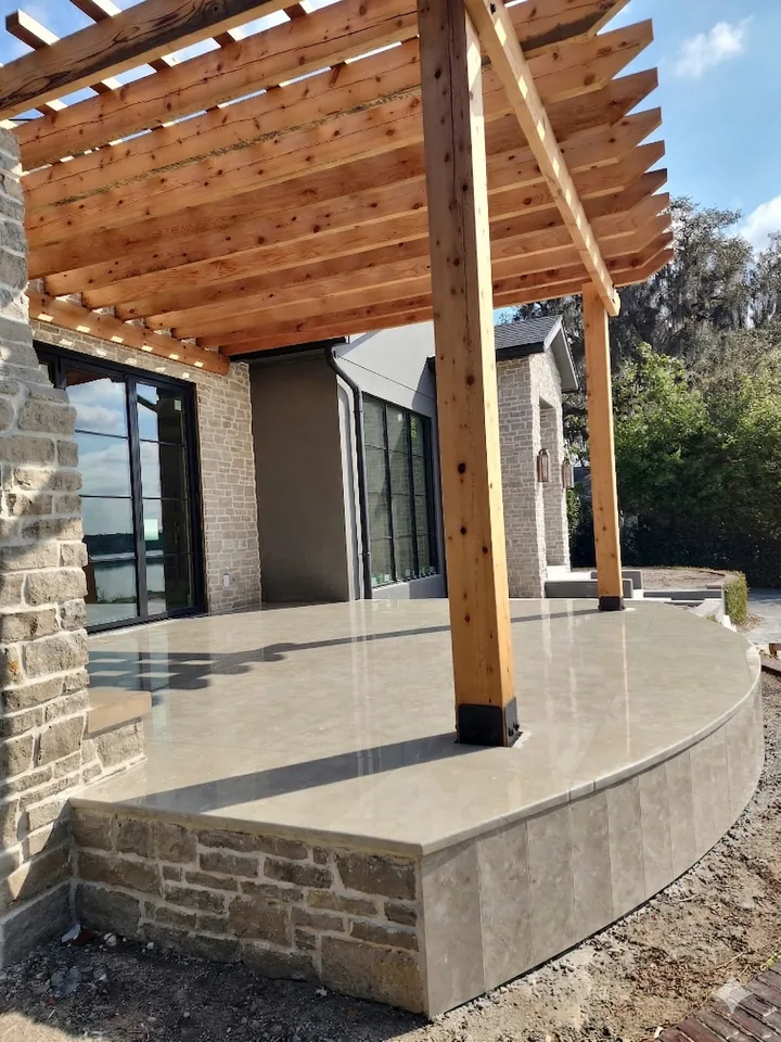 Curved raised terrace with a stone-veneer wall face and a cedar pergola, built off the rear of a lakefront home during construction
