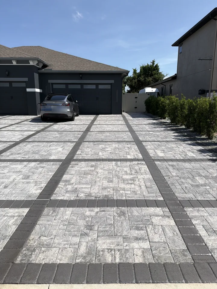 Gray paver driveway with charcoal cross bands running up to a dark two-car garage, with a matching side walkway along the hedge