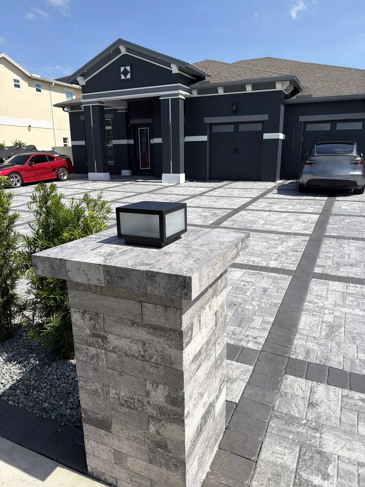 Paver-clad entry pillar with a mounted light fixture at the edge of a gray and charcoal paver driveway