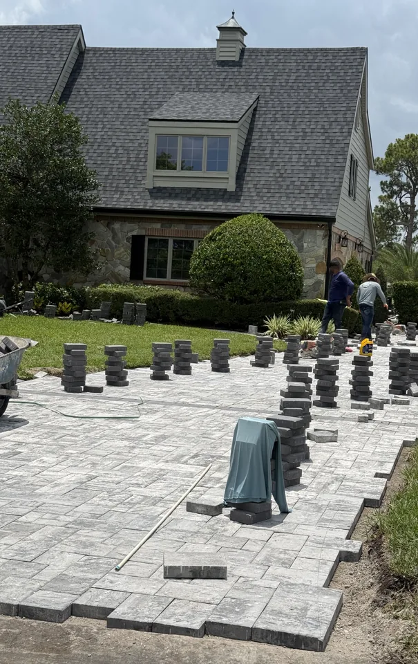 Paver driveway installation in progress: stacks of gray pavers staged on the screeded bedding sand while the crew sets the field