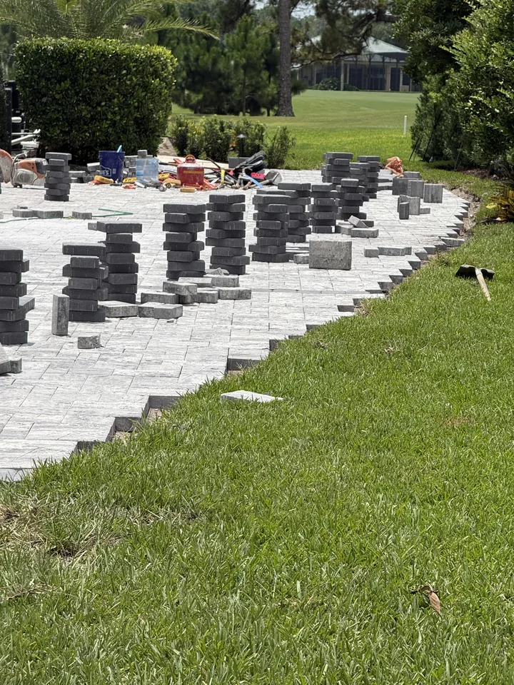Stacks of pavers lined up along the lawn edge of a driveway during installation, showing the screeded bedding layer