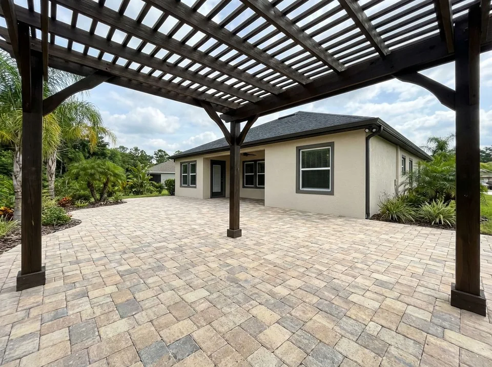 Tumbled tan-blend paver patio under a dark-stained wood pergola behind a single-story stucco home