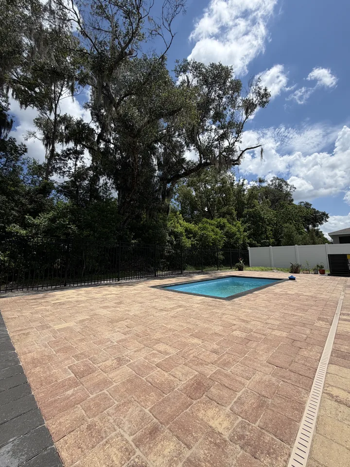 Tumbled travertine-look paver pool deck with a drain channel around a rectangular pool, shaded by mature oaks