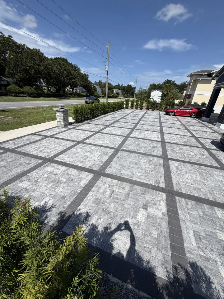 Wide gray paver driveway with charcoal grid banding and border, looking from the garage toward the street and the paver entry pillar